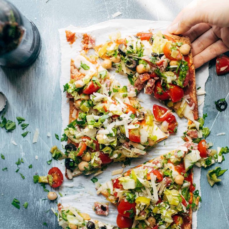 Olives, chickpeas, tomatoes, greens, herbs, and cheese on a thin crust flatbread pizza on a white plate.